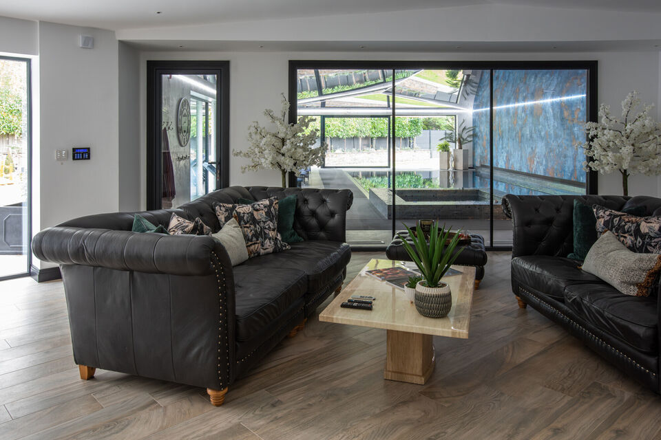 Modern pool lounge in a Cheshire residence featuring black leather Chesterfield sofas, timber flooring, and floor-to-ceiling glass doors overlooking an indoor swimming pool with a reflective stretch ceiling