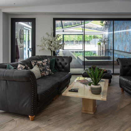 Modern pool lounge in a Cheshire residence featuring black leather Chesterfield sofas, timber flooring, and floor-to-ceiling glass doors overlooking an indoor swimming pool with a reflective stretch ceiling