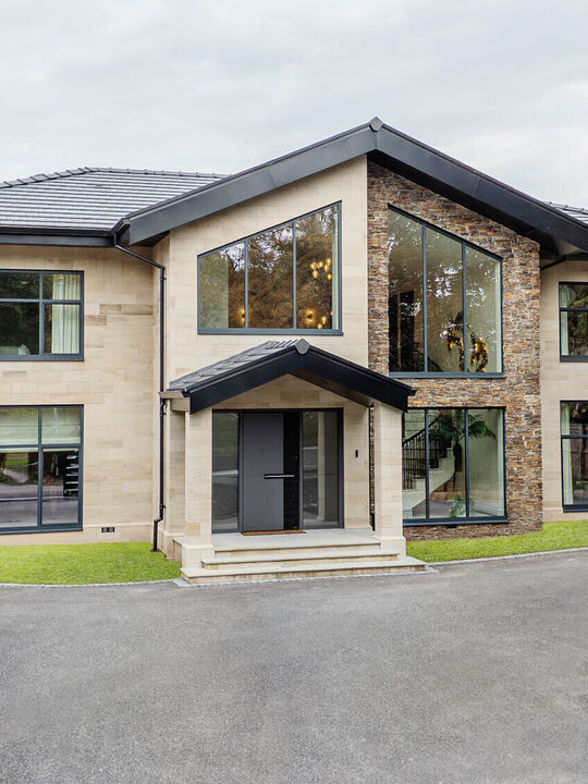 Front elevation of a bespoke Cheshire residence featuring light ashlar stone masonry, dark grey window frames, and a modern gabled entrance.
