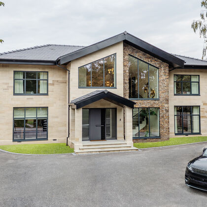Front elevation of a bespoke Cheshire residence featuring light ashlar stone masonry, dark grey window frames, and a modern gabled entrance.